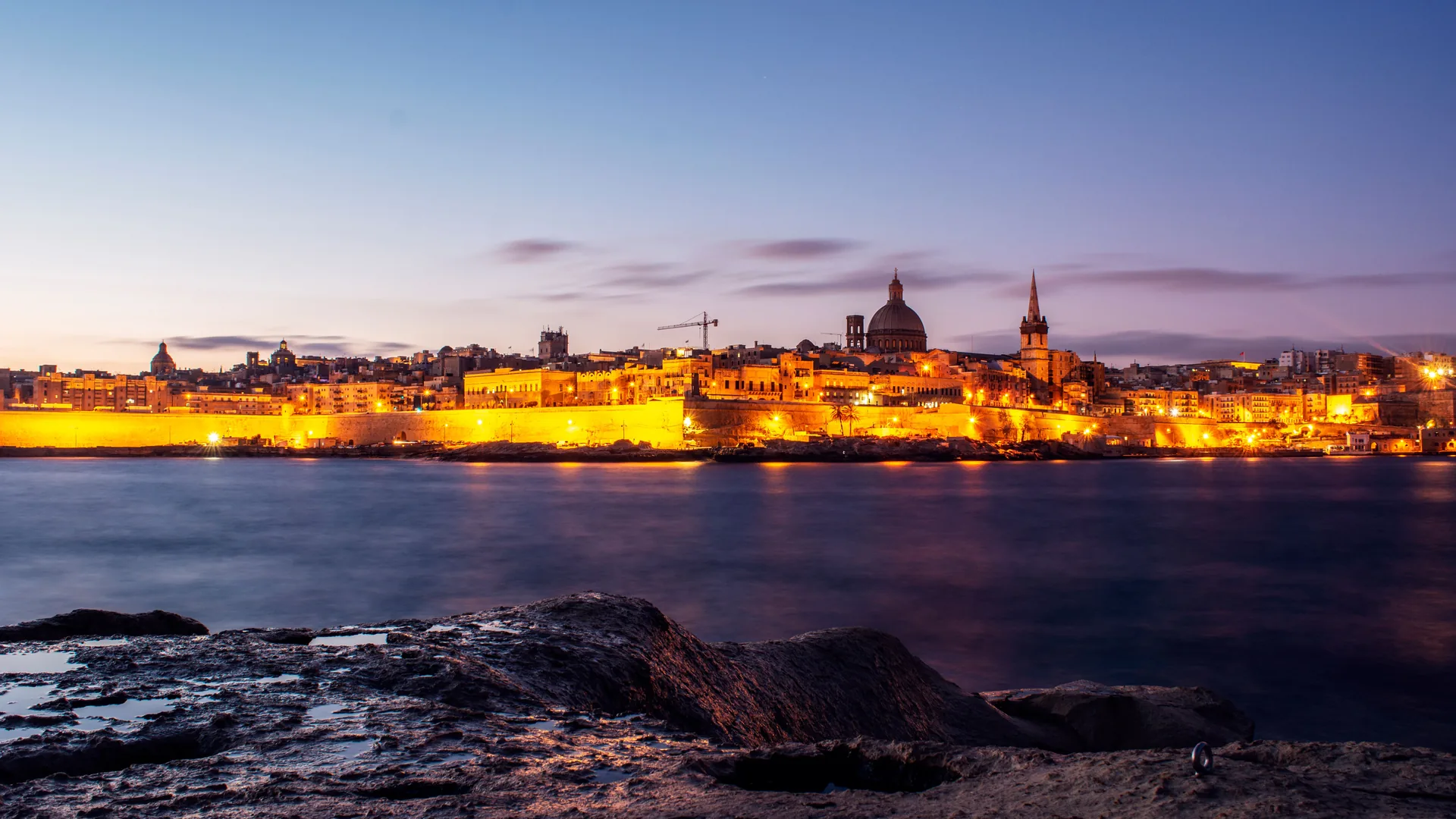 A Maltese town glowing at dusk, lights reflecting over the water — representative of a Malta evening